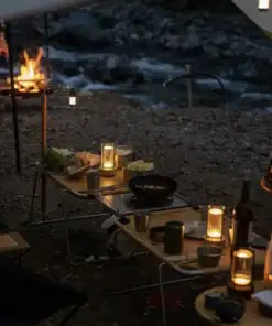 A campfire burns near a river; a table with food, drinks, and lanterns is set for an outdoor meal at dusk. | 28Alchemy