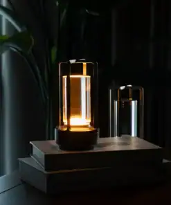 A modern lantern-style lamp glows warmly atop stacked books, with plants and curtains in the background. | 28Alchemy