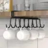 Five white cups hang from a black rack under a wooden shelf with plates and glasses on top. | 28Alchemy