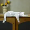 A geometric white cat sculpture lies stretched out on top of a wooden cabinet, with flowers in a vase in the background. | 28Alchemy