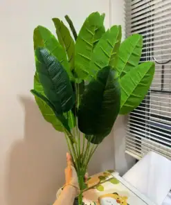 A hand holds a bunch of large, green artificial leaves near a window with blinds and a table with a cartoon animal design. | 28Alchemy