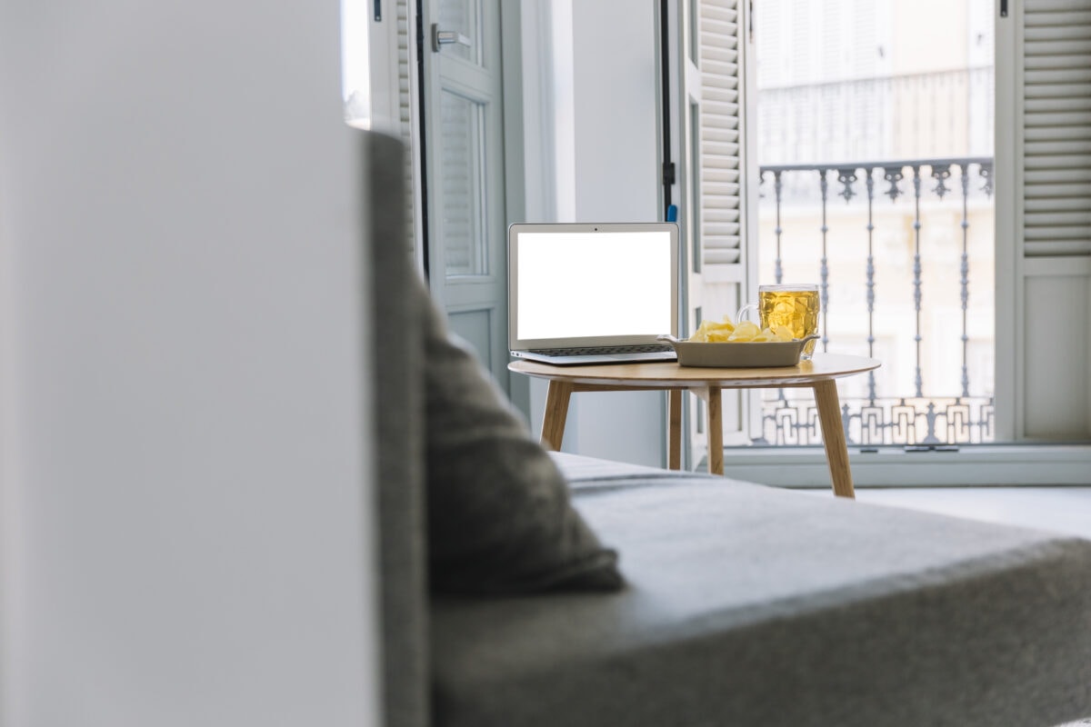 A laptop, a drink, and snacks on a wooden table next to a sofa by a window with open shutters. | 28Alchemy