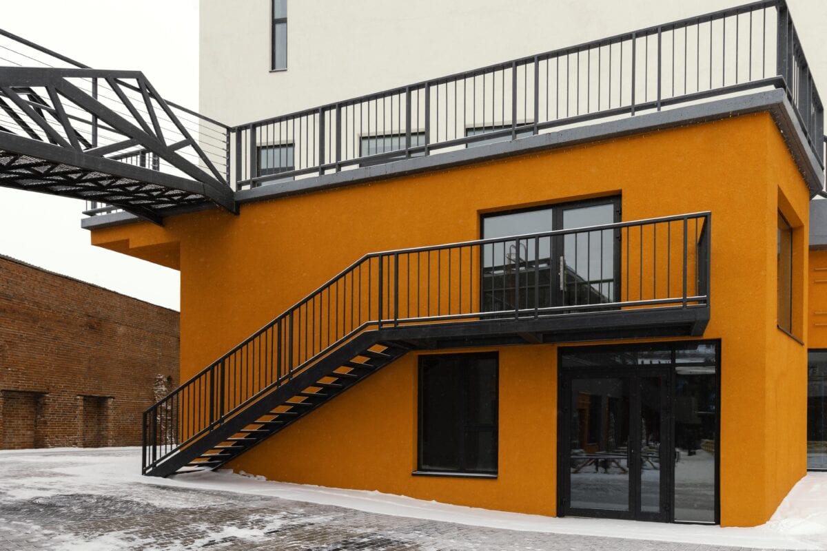 Modern orange building with black metal stairs, balcony, and a walkway, surrounded by snow and brick pavement. | 28Alchemy