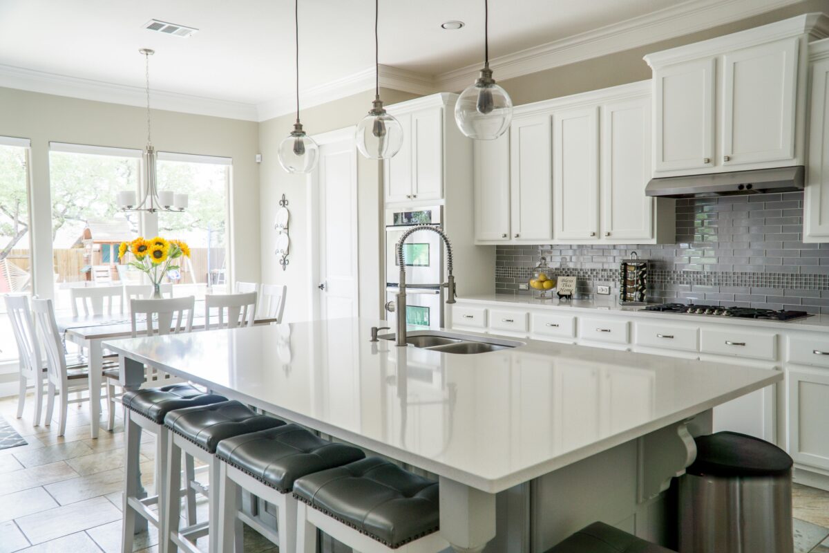 Modern white kitchen with a large island, pendant lights, and a dining area with sunflowers on the table. | 28Alchemy