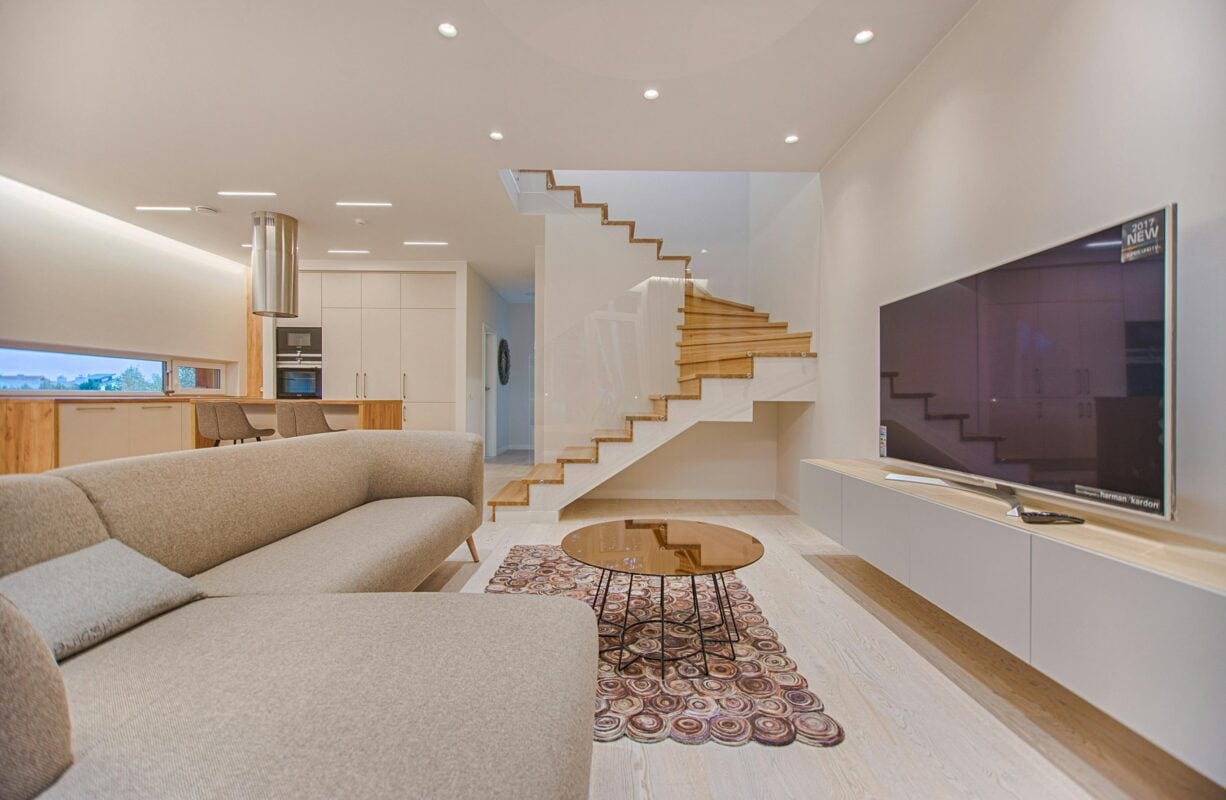 Modern living room with beige sofa, round glass coffee table, wall-mounted TV, and floating wooden staircase. | 28Alchemy