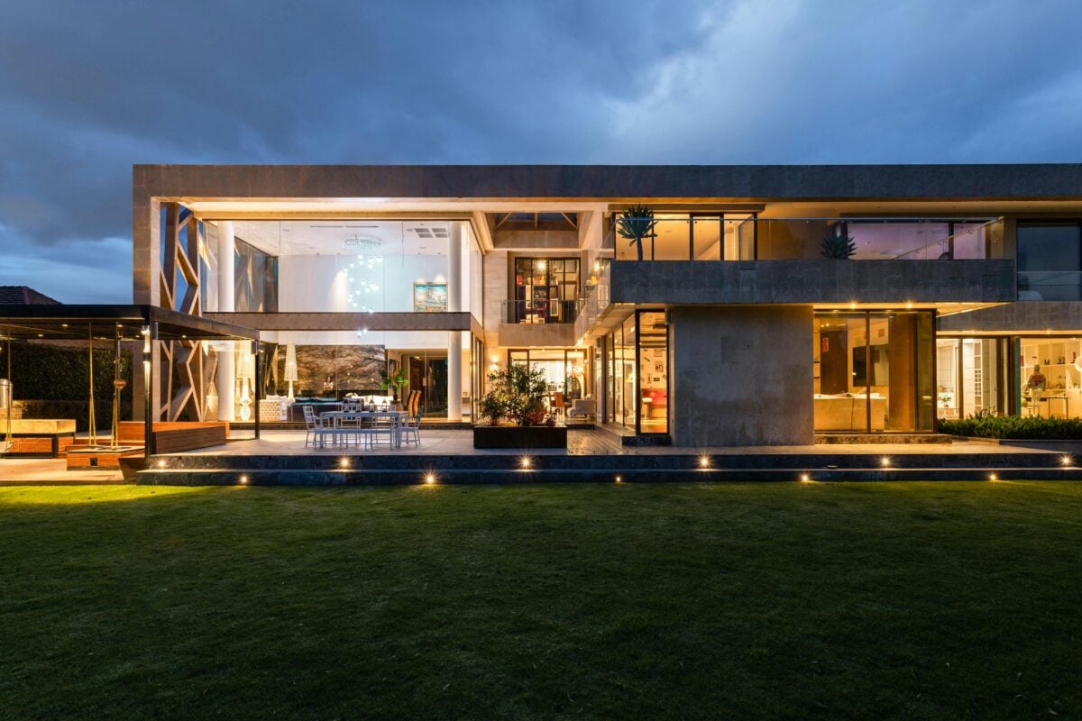 Modern two-story house with large glass windows, warm interior lighting, and a grassy lawn at dusk. | 28Alchemy
