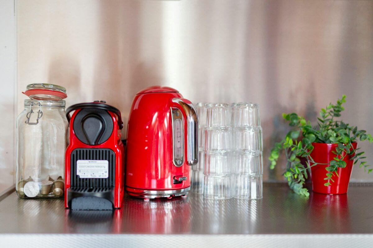 A red coffee maker and kettle sit on a counter with glass jars, stacked glasses, and a potted plant. | 28Alchemy