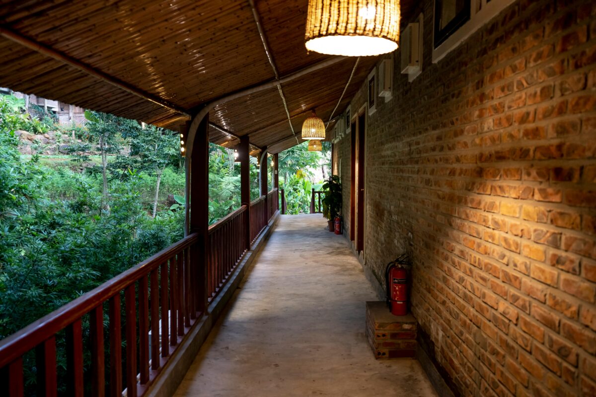 Covered corridor with brick wall, bamboo ceiling, hanging lights, and greenery visible outside the railing. | 28Alchemy