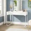 White vanity table with a mirror framed by lights, makeup items, and a vase with flowers against a blue wall. | 28Alchemy