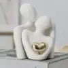 Minimalist white sculpture of two faceless figures embracing, with a gold heart in the center. | 28Alchemy
