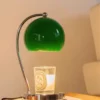 A green desk lamp illuminates a candle placed on a book atop a wooden surface next to a beige sofa. | 28Alchemy