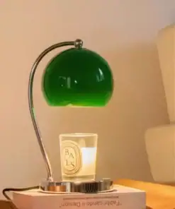A green desk lamp illuminates a candle placed on a book atop a wooden surface next to a beige sofa. | 28Alchemy