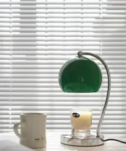 A green desk lamp, a lit candle, and a white mug sit on a table in front of closed window blinds. | 28Alchemy