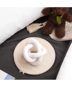 A white knot-shaped pillow on a round mat beside a teddy bear with a green ribbon on a bed. | 28Alchemy