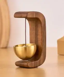A small brass bell hanging by a cord from a curved wooden stand on a light wood surface. | 28Alchemy