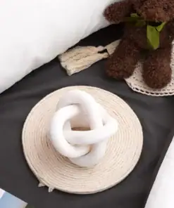 A white knotted sculpture sits on a woven mat near a brown teddy bear on a dark bedsheet. | 28Alchemy