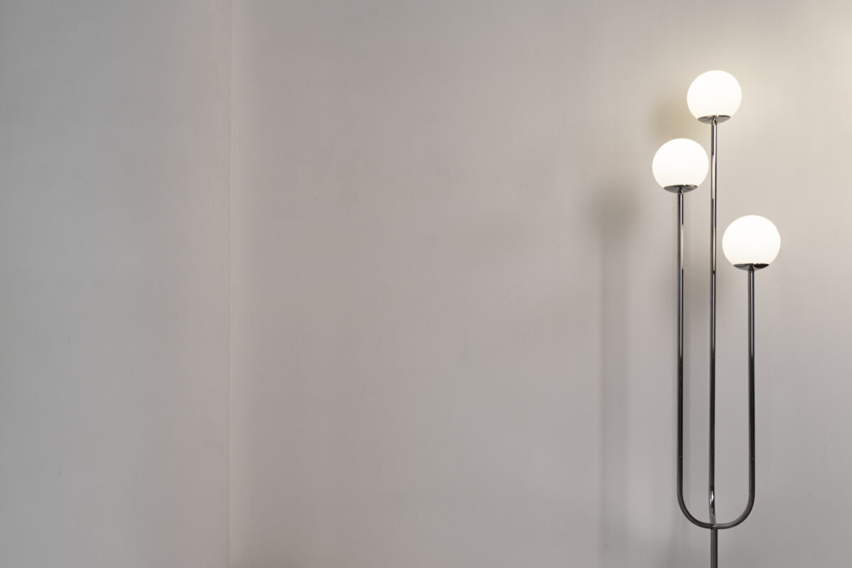 A modern floor lamp with three glowing round bulbs stands against a plain, light-colored wall. | 28Alchemy