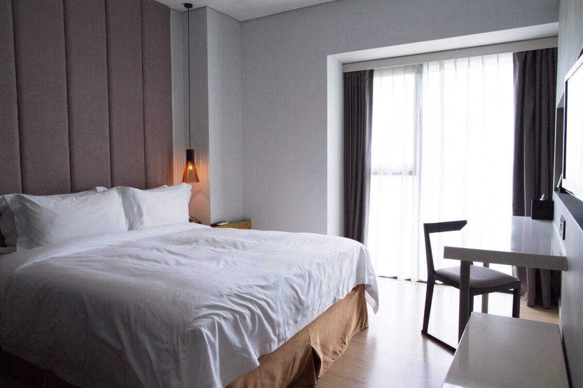 Modern hotel room with a large bed, desk, chair, and window with sheer curtains letting in natural light. | 28Alchemy