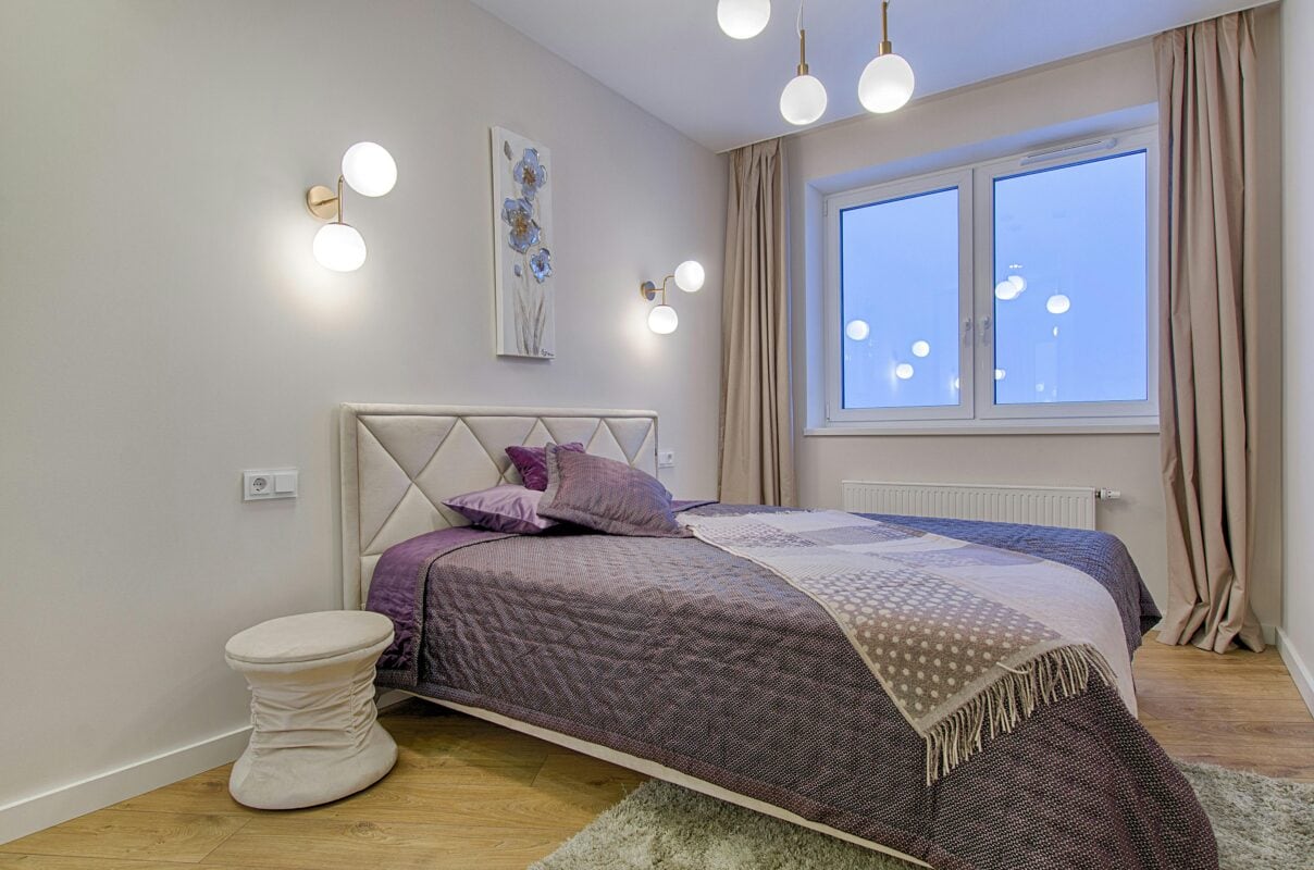 Modern bedroom with a double bed, purple bedding, house lighting, artwork, and large window with beige curtains. | 28Alchemy