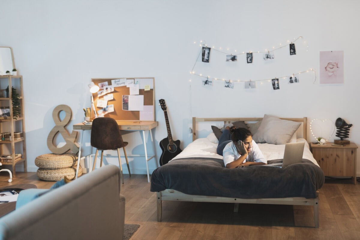 Person lying on bed using a laptop in a cozy bedroom with warm lights, a desk, guitar, and photo garland on the wall. | 28Alchemy