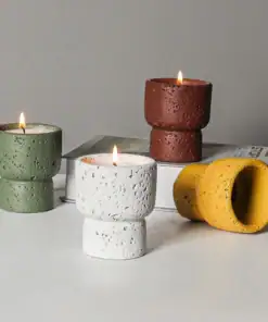 Four textured candles in green, white, brown, and yellow holders, three lit, sit on a white surface by a book. | 28Alchemy