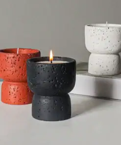 Three textured candles in modern, hourglass-shaped holders: one black (lit), one red, and one white, on a table. | 28Alchemy