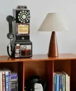 A vintage rotary payphone and a table lamp sit on a wooden shelf next to books and a small black-and-white figurine. | 28Alchemy