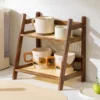 Two-tier wooden shelf holding four ceramic mugs and two small bowls, displayed on a kitchen counter in sunlight. | 28Alchemy