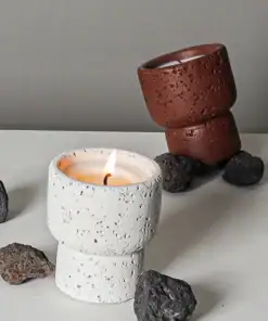 A lit white candle in a textured holder sits among rocks; a similar empty brown holder stands behind it. | 28Alchemy