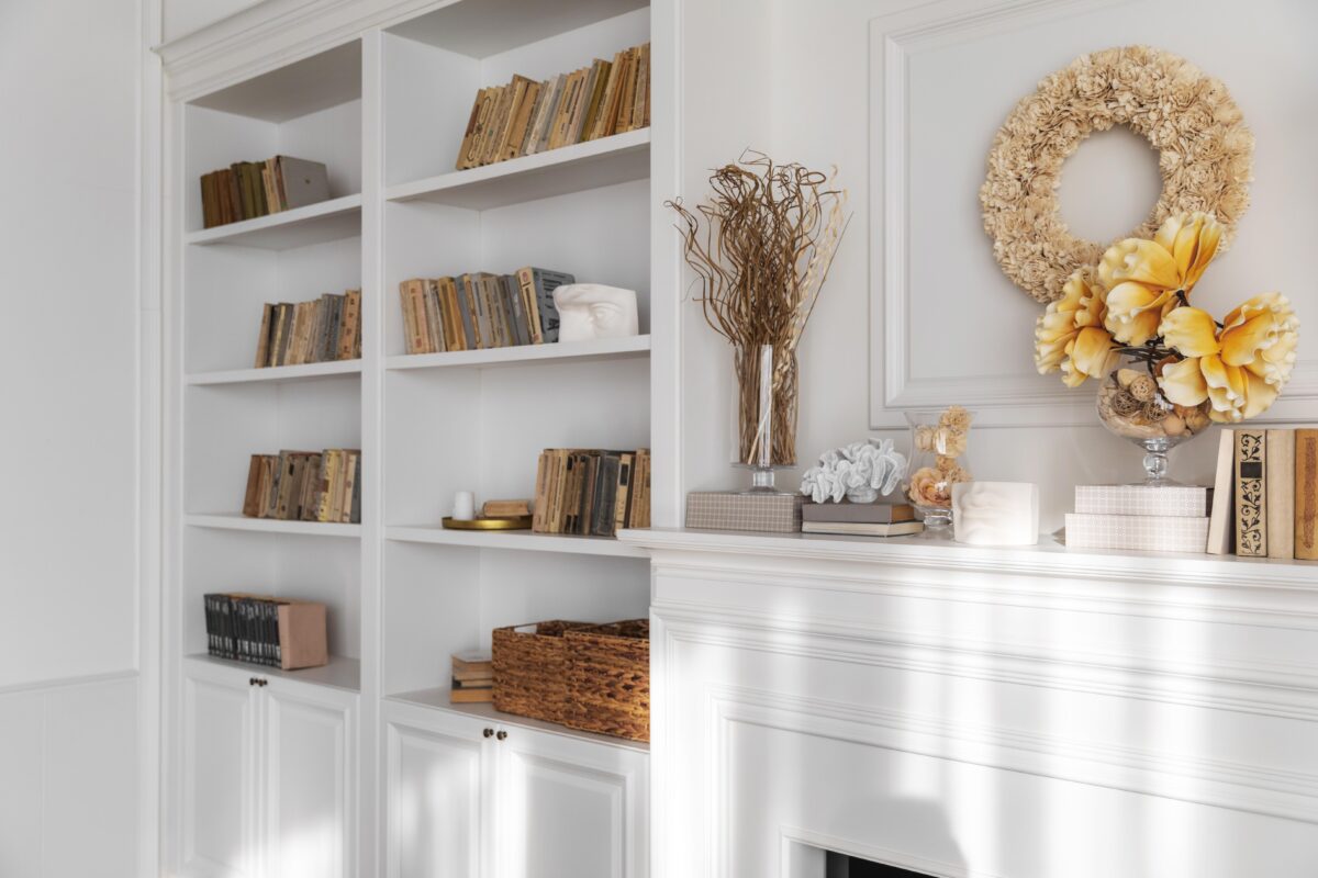 White shelves with vintage books, wicker baskets, and decor beside a white mantel with flowers, wreath, and decorative boxes. | 28Alchemy