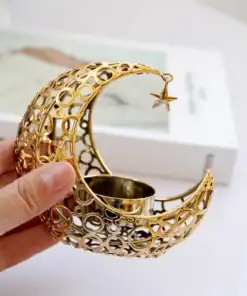 A hand holds a gold crescent moon-shaped candle holder with a hanging star, next to a closed book. | 28Alchemy