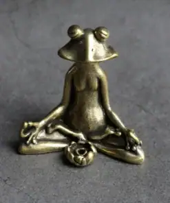 Small brass frog figurine sitting cross-legged in a meditative yoga pose on a gray surface. | 28Alchemy