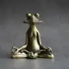 A bronze frog figurine sits in a meditative pose with crossed legs and closed eyes on a gray surface. | 28Alchemy