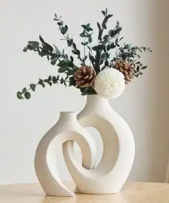 Two modern cream vases with a curved design hold eucalyptus, pinecones, and a white flower on a wooden table. | 28Alchemy