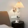 Wall lamp above a bedside table with a teacup, saucer, hourglass, and power outlets on a light-colored wall. | 28Alchemy