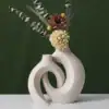 Two modern cream-colored abstract vases with dried flowers and foliage against a dark green background. | 28Alchemy