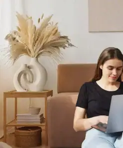 A woman sits on a beige couch using a laptop, with pampas grass in vases on a wooden side table beside her. | 28Alchemy