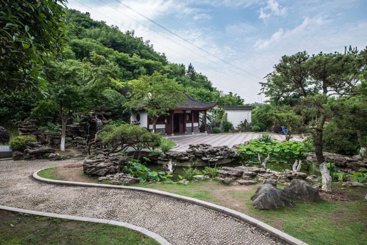 A serene Chinese garden with a pond, stone path, lush greenery, and a traditional pavilion under a partly cloudy sky. | 28Alchemy