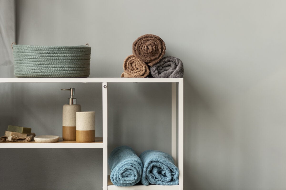 White shelf with blue, brown, and gray rolled towels, a basket, and soap dispensers against a gray wall. | 28Alchemy