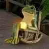 A frog figurine relaxes on a rocking chair, holding a glowing candle, with plants and a wooden deck in the background. | 28Alchemy