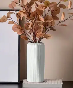 Tall white ribbed vase with brown dried branches, placed on a closed book beside a picture frame against a beige wall. | 28Alchemy