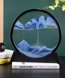A circular sand art frame with blue sand forming mountain shapes, displayed on books with a plant and radio nearby. | 28Alchemy