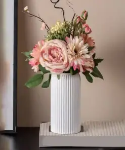 A white ribbed vase holds pink and cream flowers with green leaves, placed on a closed book on a dark surface. | 28Alchemy