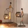 Two metal candle holders shaped like figures holding lanterns, with lit candles inside, placed on a table near books. | 28Alchemy