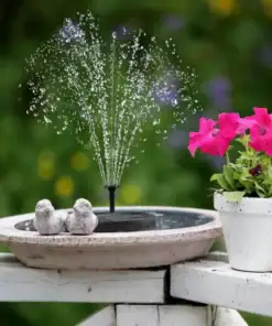 A small birdbath fountain sprays water beside a potted plant with bright pink flowers on a white wooden surface. | 28Alchemy