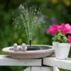 Solar fountain spraying water in a birdbath with two bird figurines, next to a pot of vibrant pink flowers. | 28Alchemy