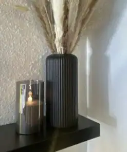 A black ribbed vase with dried grasses next to a lit candle in a glass holder on a black shelf. | 28Alchemy