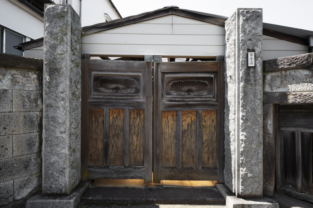 A traditional wooden Japanese gate with stone pillars and weathered wood panels, closed under a simple roof. | 28Alchemy