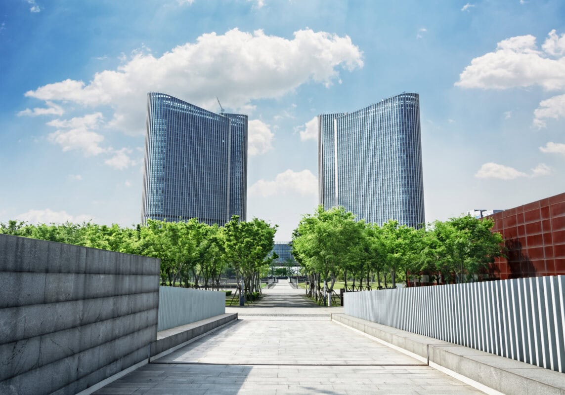 Two modern glass skyscrapers rise behind a row of green trees under a blue sky with clouds. | 28Alchemy
