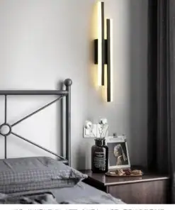 Modern vertical wall sconce emits soft light beside a bed with gray bedding and a nightstand holding decor and a framed photo. | 28Alchemy